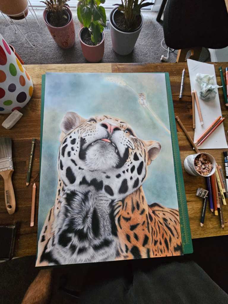 A close-up artwork of a jaguar looking up, featuring intricate details in its fur and facial expression, set on a wooden table surrounded by art supplies like pencils, a brush, and plant pots in the background.